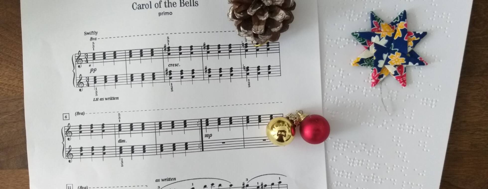 Ein Notenblatt in Schwarzdruck und Braille von "Carol of the Bells" mit einem Tannenzapfen, Glöckchen und einen Weihnachtsstern