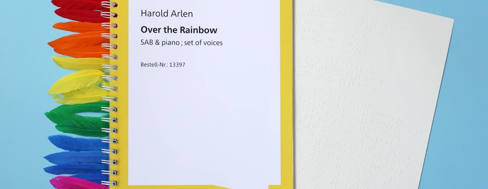 Ein Ringbuch, aus dem eine Braillenotenseite ragt vor hellblauem Hintergrund. Links schauen Federn in den Farben des Regenbogens aus dem Buch. Text: Harold Arlen. Over the Rainbow. SAB und piano, set of voices