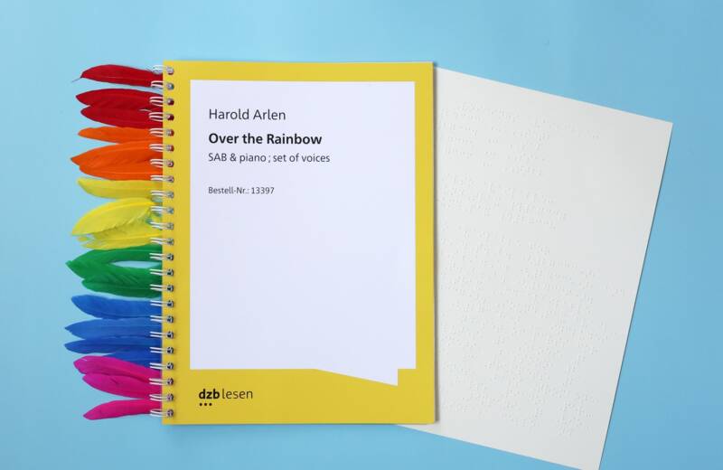 Ein Ringbuch, aus dem eine Braillenotenseite ragt vor hellblauem Hintergrund. Links schauen Federn in den Farben des Regenbogens aus dem Buch. Text: Harold Arlen. Over the Rainbow. SAB und piano, set of voices